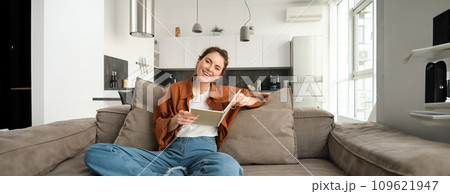 Young woman sits at home on sofa, reading her notes, holding notebook, studying for exam, student revising in her living room 109621947