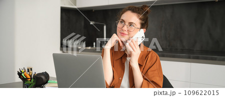Portrait of young self-employed woman, entrepreneur working from home, freelancer calling client. Girl making an order, talking to someone on phone, sitting in kitchen with laptop 109622015