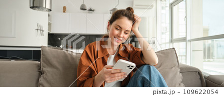 Portrait of happy young brunette woman sitting on sofa with smartphone, reading notification, messaging on mobile phone, resting on couch at home, smiling while looking at device screen Portrait of happy young brunette woman sitting on sofa with smartphone, reading notification, messaging on mobile phone, resting on couch at home, smiling while looking at device screen 109622054