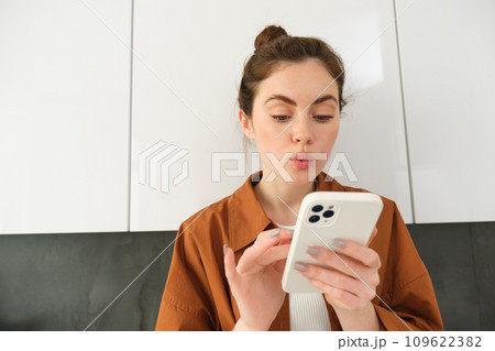 Portrait of young woman at home, looking with interest at smartphone screen, concept of online shopping, buying on mobile apps, picking something using application on phone Portrait of young woman at home, looking with interest at smartphone screen, concept of online shopping, buying on mobile apps, picking something using application on phone 109622382