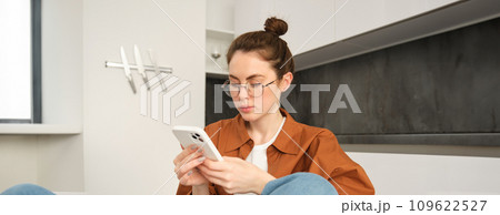 Portrait of young brunette woman in glasses, sitting at home with mobile phone, using her smartphone and smiling, reading message Portrait of young brunette woman in glasses, sitting at home with mobile phone, using her smartphone and smiling, reading message 109622527