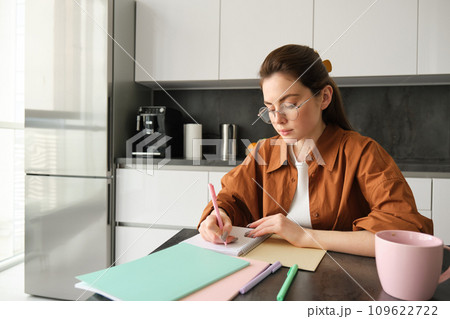 Portrait of woman looking concentrated, writing down notes, doing homework in kitchen, drinking coffee, studying at home 109622722