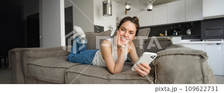 Portrait of woman spending time at home, resting on couch in living room, holding smartphone, smiling and looking happy at camera 109622731