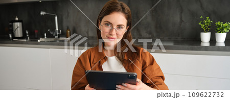 Portrait of brunette woman in glasses, sitting in kitchen at home, holding digital tablet, reading on gadget 109622732