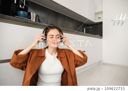 Cute brunette woman on kitchen floor, wears headphones, listens music and smiles, enjoys sound quality 109622733
