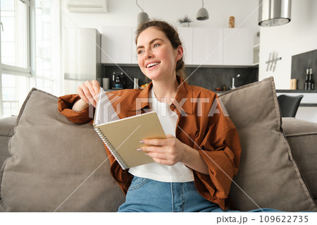 Portrait of young smiling beautiful woman, sitting on sofa in her living room, holding study material, doing homework, reading notes in notebook 109622735