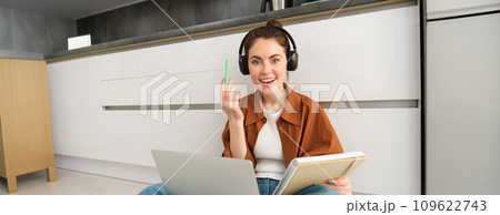 Portrait of young woman, modern student sits at home on floor and doing homework on laptop, listens music in headphones while making assignment or project for college 109622743