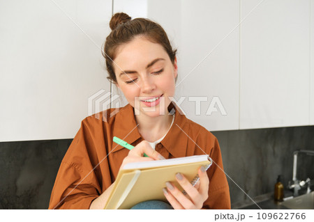 Portrait of cute young woman with notebook, sitting in the kitchen and writing her to do list, making tasks for week, puts notes in schedule or planner Portrait of cute young woman with notebook, sitting in the kitchen and writing her to do list, making tasks for week, puts notes in schedule or planner 109622766