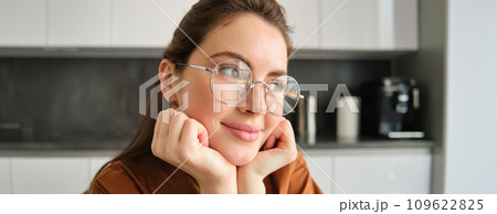 Portrait of lovely, happy young woman, wearing glasses, sitting in kitchen with dreamy, thoughtful face expression, spending time at home Portrait of lovely, happy young woman, wearing glasses, sitting in kitchen with dreamy, thoughtful face expression, spending time at home 109622825