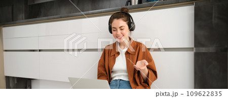 Portrait of young smiling woman explains something over the video call, sits in headphones with laptop, works from home 109622835