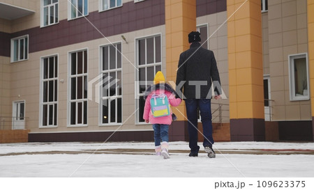 father takes child school holding his hand. happy family. snowy road school. father leads his little girl with school backpack through snow school. cold season. Dad holds hand child kid with backpack. 109623375