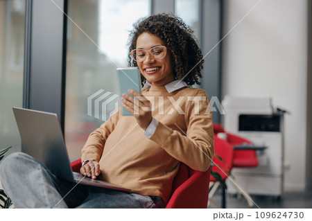 Smiling female manager is use phone while working on laptop in office near window Smiling female manager is use phone while working on laptop in office near window 109624730