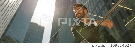 Smiling businessman in suit is standing with laptop on office terrace and looks away 109624927