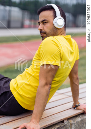man athlete, wearing wireless headphones resting on wooden bench, taking a break after a workout at the sports arena. 109625305