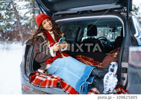 Young woman sitting on car trunk, sending messages in sunny winter forest. Lifestyle concept. 109625805