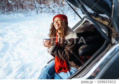 Happy woman holds a thermos and drinks tea sitting in car trunk in winter forest. Lifestyle concept. Happy woman holds a thermos and drinks tea sitting in car trunk in winter forest. Lifestyle concept. 109625812