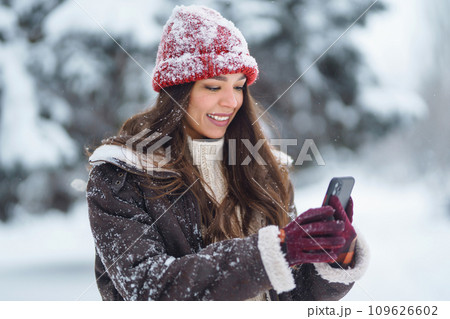 Happy woman in a red hat with a phone takes a selfie, blogs in the winter forest. Concept of nature. Happy woman in a red hat with a phone takes a selfie, blogs in the winter forest. Concept of nature. 109626602
