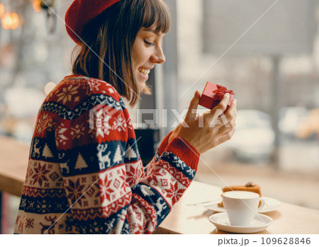 Joyful woman in a festive sweater opening a gift at a cozy cafe window Joyful woman in a festive sweater opening a gift at a cozy cafe window 109628846
