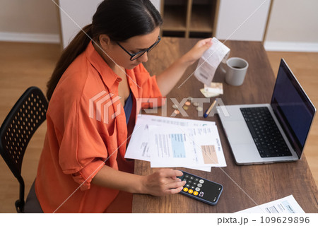 top view Desk Finance: A woman, with a laptop on her desk, tracks household expenses using her smartphone calculator. 109629896