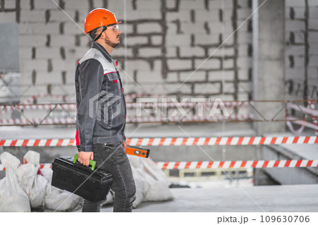 A worker in overalls and a helmet came to a new work site. Builder in uniform carrying toolbox at construction site, copy space 109630706