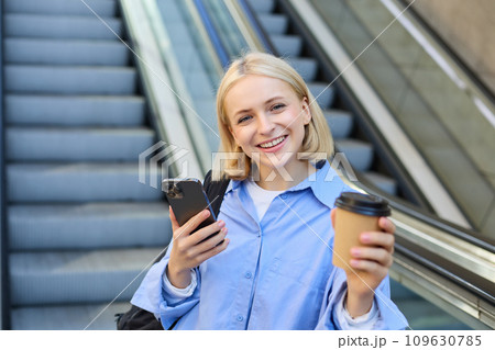 Portrait of young modern woman with backpack, smartphone, drinking cup of coffee, standing near escalator and smiling at camera Portrait of young modern woman with backpack, smartphone, drinking cup of coffee, standing near escalator and smiling at camera 109630785