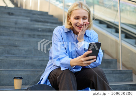 Portrait of young blond woman, sitting outside campus on city stairs, looking at smartphone with happy surprised face, receive positive good news on video chat, watching interesting video Portrait of young blond woman, sitting outside campus on city stairs, looking at smartphone with happy surprised face, receive positive good news on video chat, watching interesting video 109630806