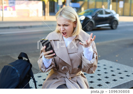 Portrait of angry young woman, shouting at her mobile phone in frustrated, being outraged, sitting on street bench outdoors Portrait of angry young woman, shouting at her mobile phone in frustrated, being outraged, sitting on street bench outdoors 109630808