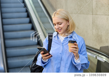 Portrait of blond young woman, reading message on her mobile phone, drinking takeaway coffee, standing near escalator with backpack 109630809