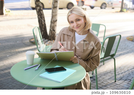 Image of young smiling woman, sitting in outdoor cafe, doing sketches, drawing art on street, spending time on coffee shop and creating art Image of young smiling woman, sitting in outdoor cafe, doing sketches, drawing art on street, spending time on coffee shop and creating art 109630810