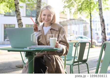 Portrait of young smiling blond woman, working on laptop, sitting in outdoor cafe on street, drinking coffee Portrait of young smiling blond woman, working on laptop, sitting in outdoor cafe on street, drinking coffee 109630811