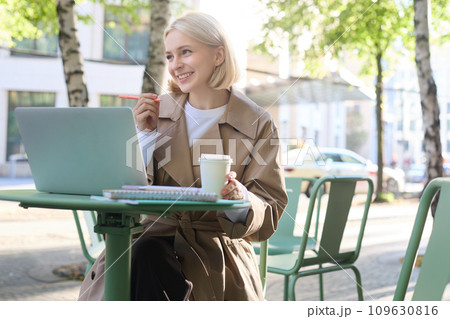 Image of young blond modern woman, sitting with laptop outside in cafe, drinking coffee drink and working on project, using computer 109630816