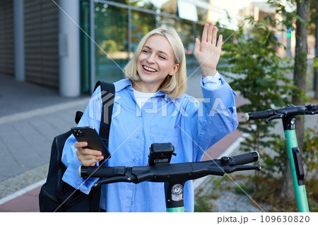 Young cheerful woman, tried to scan QR code on electric scooter on street, using mobile phone to rent it, saying hello to friend and waving hand at someone 109630820