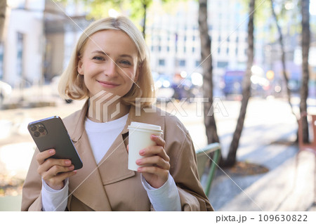 Portrait of cute blond woman with smartphone, drinking chai, coffee at favourite place in city centre, sitting outdoors, holding mobile phone 109630822