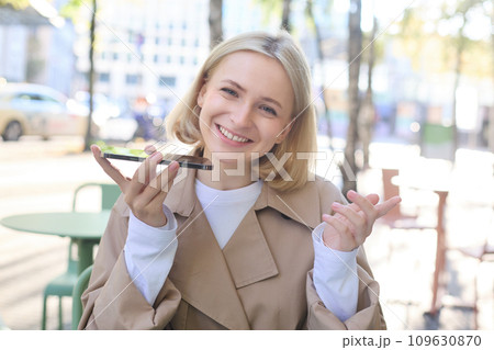 Close up portrait of smiling blond woman, girl with mobile phone outdoors, sitting in cafe, recording voice message, talking into smartphone with microphone near mouth 109630870
