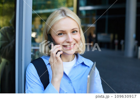 Close up portrait of modern young woman, student standing near building in city centre, talking on mobile phone, having chat on smartphone and smiling Close up portrait of modern young woman, student standing near building in city centre, talking on mobile phone, having chat on smartphone and smiling 109630917