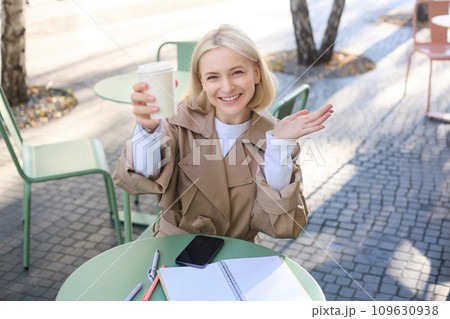 Food and drink concept. Young happy woman sitting in cafe, enjoying bright day outdoors, raising cup of coffee and smiling, drinking chai 109630938