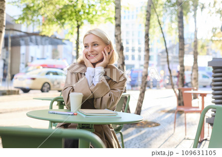 Urban lifestyle concept. Young beautiful woman sitting in outdoor cafe, contemplating city life, drinking coffee and smiling on street 109631105