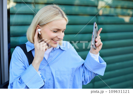 Close up portrait of blond smiling female model, college student in earphones, listening to music on street, holding smartphone, waiting for someone outdoors 109631121