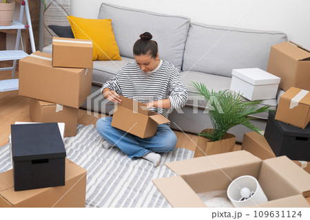 Life in Motion: Young woman unfolds her new chapter, encircled by carton boxes in her home, reflecting the choices of relocation, rentals, or home buying Life in Motion: Young woman unfolds her new chapter, encircled by carton boxes in her home, reflecting the choices of relocation, rentals, or home buying 109631124