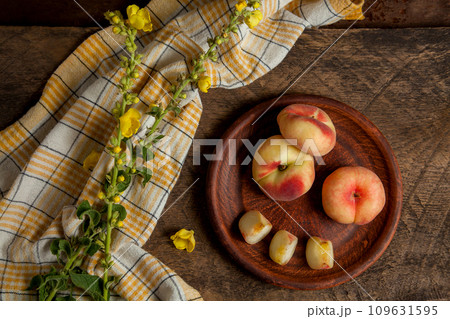Clay plate with cut and whole saturn peaches or flat peaches on wooden background with yellow kitchen towel.. Clay plate with cut and whole saturn peaches or flat peaches on wooden background with yellow kitchen towel.. 109631595