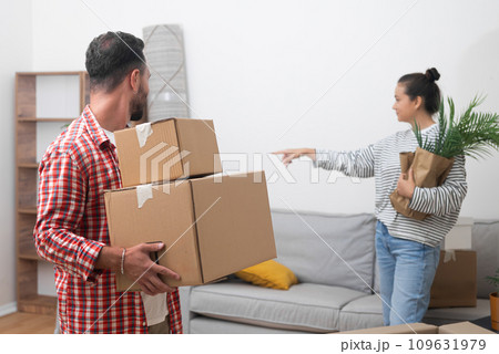 Young woman with packed house plant in hands points index finger to man with cardboard parcels to move boxes with goods moving to new place in city and relocating Young woman with packed house plant in hands points index finger to man with cardboard parcels to move boxes with goods moving to new place in city and relocating 109631979
