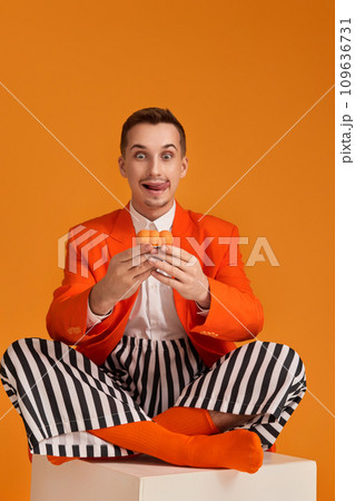 extravagant funny young man in orange jacket and striped pants holding cupcake on orange background. 109636731