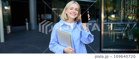 Happy smiling woman showing smartphone screen, demonstrating mobile phone display, standing in college outdoors, posing near campus with laptop and telephone Happy smiling woman showing smartphone screen, demonstrating mobile phone display, standing in college outdoors, posing near campus with laptop and telephone 109640720