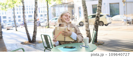 Image of young beautiful woman, student doing homework in an outdoor coffee shop, holding journal or planner, writing in notebook, smiling happily 109640727