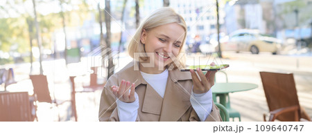 Close up portrait of beautiful young blond girl, records voice mail, holding smartphone near mouth and smiling, sitting in cafe outdoors 109640747
