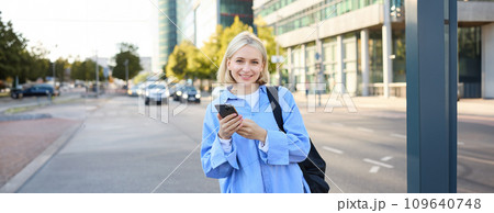 Street style shot of young smiling woman with smartphone, standing on street, bright sunny day, road with cars behind her back, using mobile phone, waiting for someone outdoors 109640748