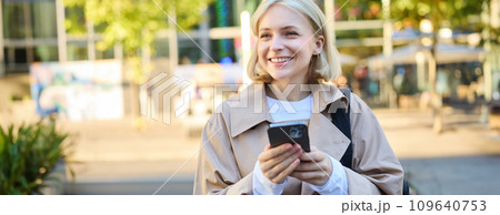 Carefree smiling woman standing on sunny street, using mobile phone, waiting for car or bus with smartphone, looking around Carefree smiling woman standing on sunny street, using mobile phone, waiting for car or bus with smartphone, looking around 109640753