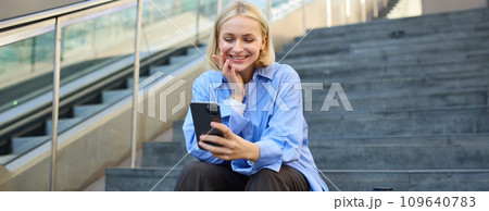 Image of young happy woman, sitting on stairs outside on street, taking selfie on smartphone camera, posing for photo social media profile, smiling 109640783