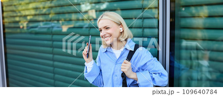 Close up portrait of blond smiling female model, college student in earphones, listening to music on street, holding smartphone, waiting for someone outdoors Close up portrait of blond smiling female model, college student in earphones, listening to music on street, holding smartphone, waiting for someone outdoors 109640784