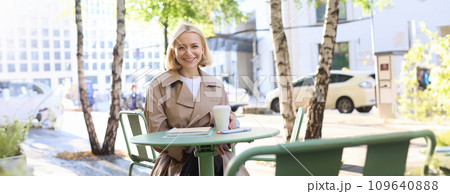 Urban lifestyle concept. Young beautiful woman sitting in outdoor cafe, contemplating city life, drinking coffee and smiling on street Urban lifestyle concept. Young beautiful woman sitting in outdoor cafe, contemplating city life, drinking coffee and smiling on street 109640888